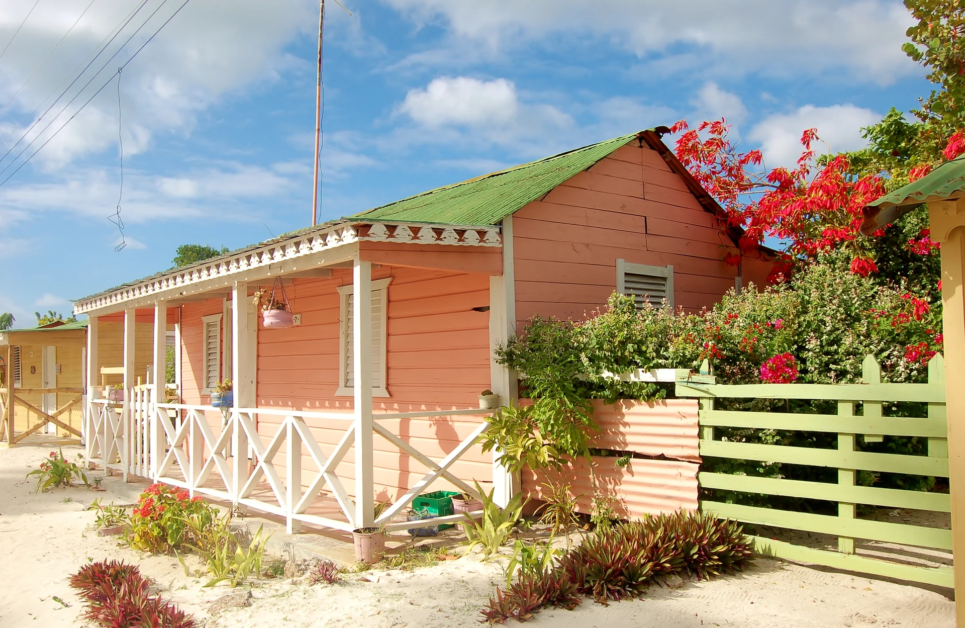 Casette di legno colorate, con una veranda bianca e arbusti in fiore, sorgono su un terreno sabbioso sull’isola di Saona.