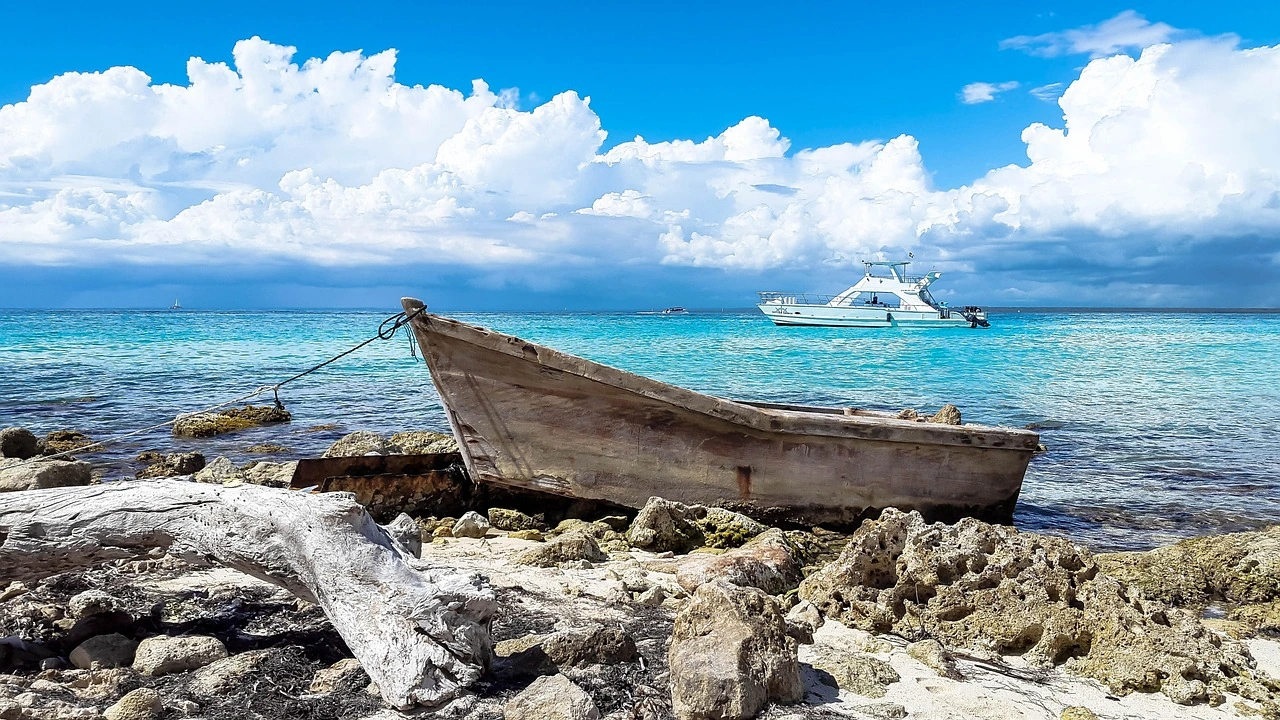 Una barca in legno segnata dalle intemperie giace sugli scogli costieri di Saona accanto all’acqua turchese, con uno yacht bianco al largo sotto nuvole chiare.