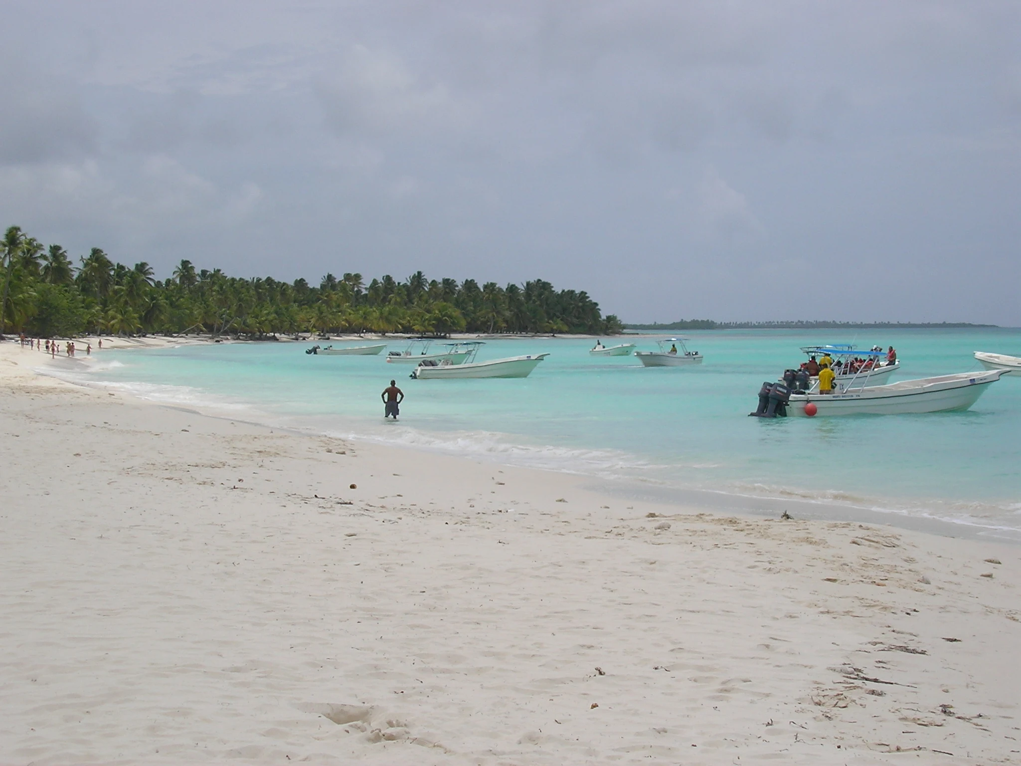 Piccoli motoscafi galleggiano accanto a un’ampia spiaggia bianca di Saona e ad acque turchesi chiare sotto un cielo nuvoloso.