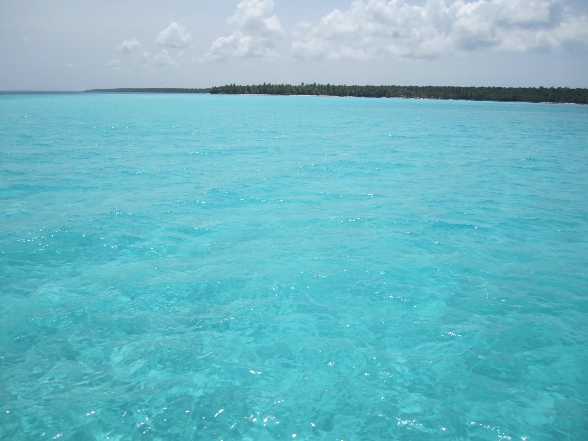 Acque basse verde-azzurre luminose si estendono verso la costa bassa e verde di Saona sotto nuvole leggere.