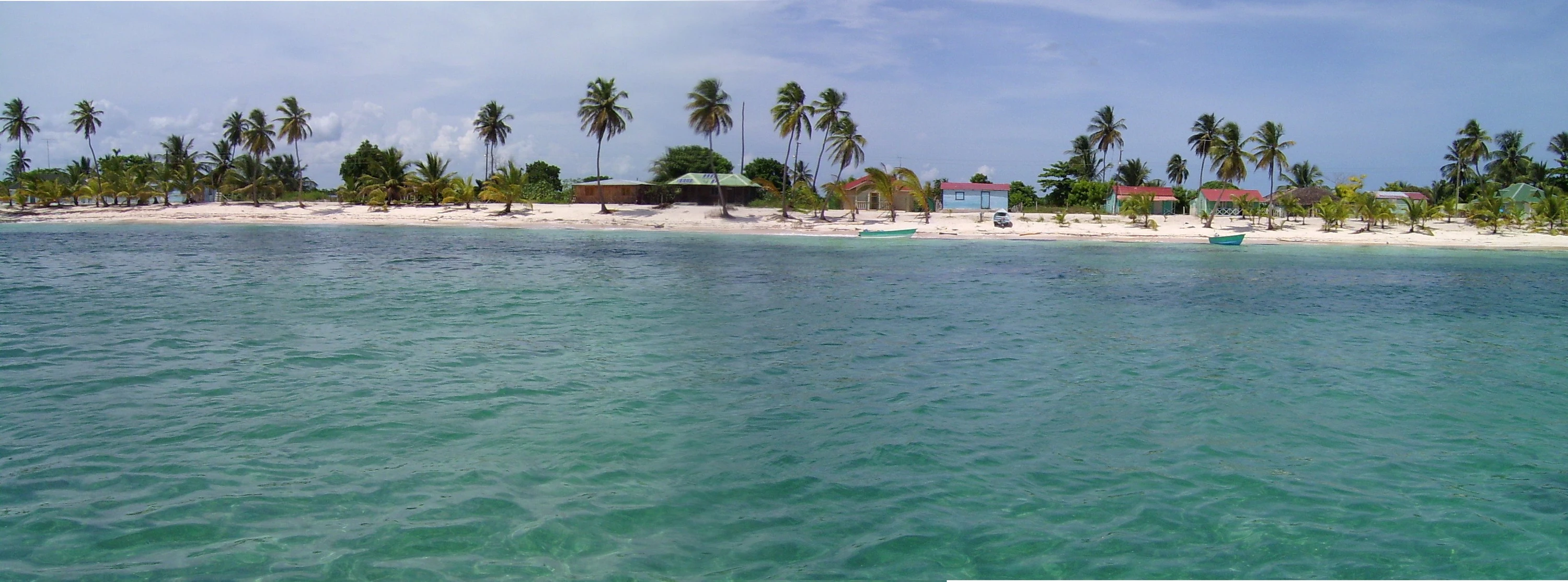 Un’ampia vista sul mare blu-verde verso una spiaggia chiara di Saona con palme da cocco e ripari sparsi.