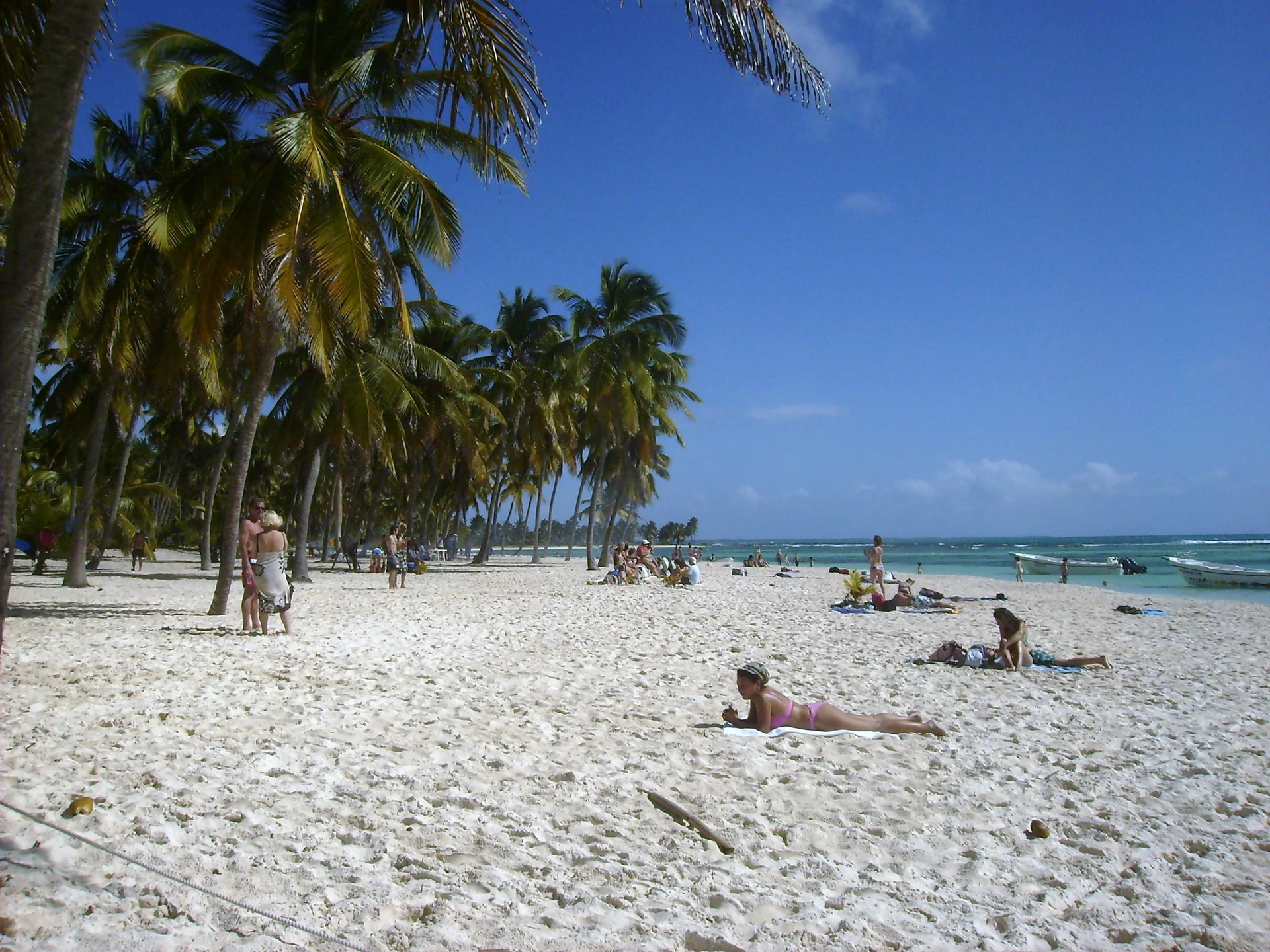 I visitatori si rilassano su un’ampia spiaggia bianca di Saona sotto le palme da cocco, accanto all’acqua turchese e a piccole barche.