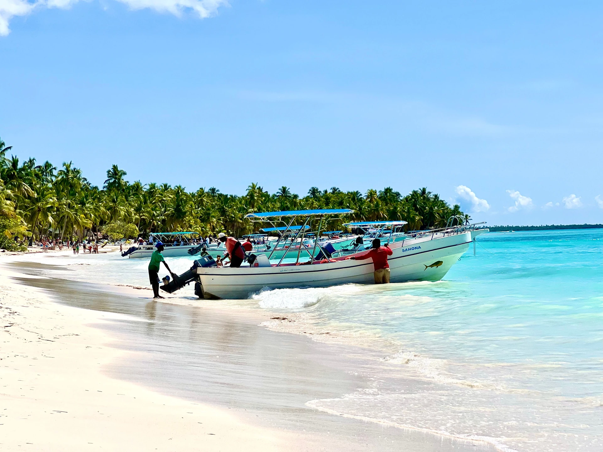 Piccole barche da escursione sono ferme su una spiaggia bianca di Saona accanto ad acqua turchese brillante e palme da cocco.