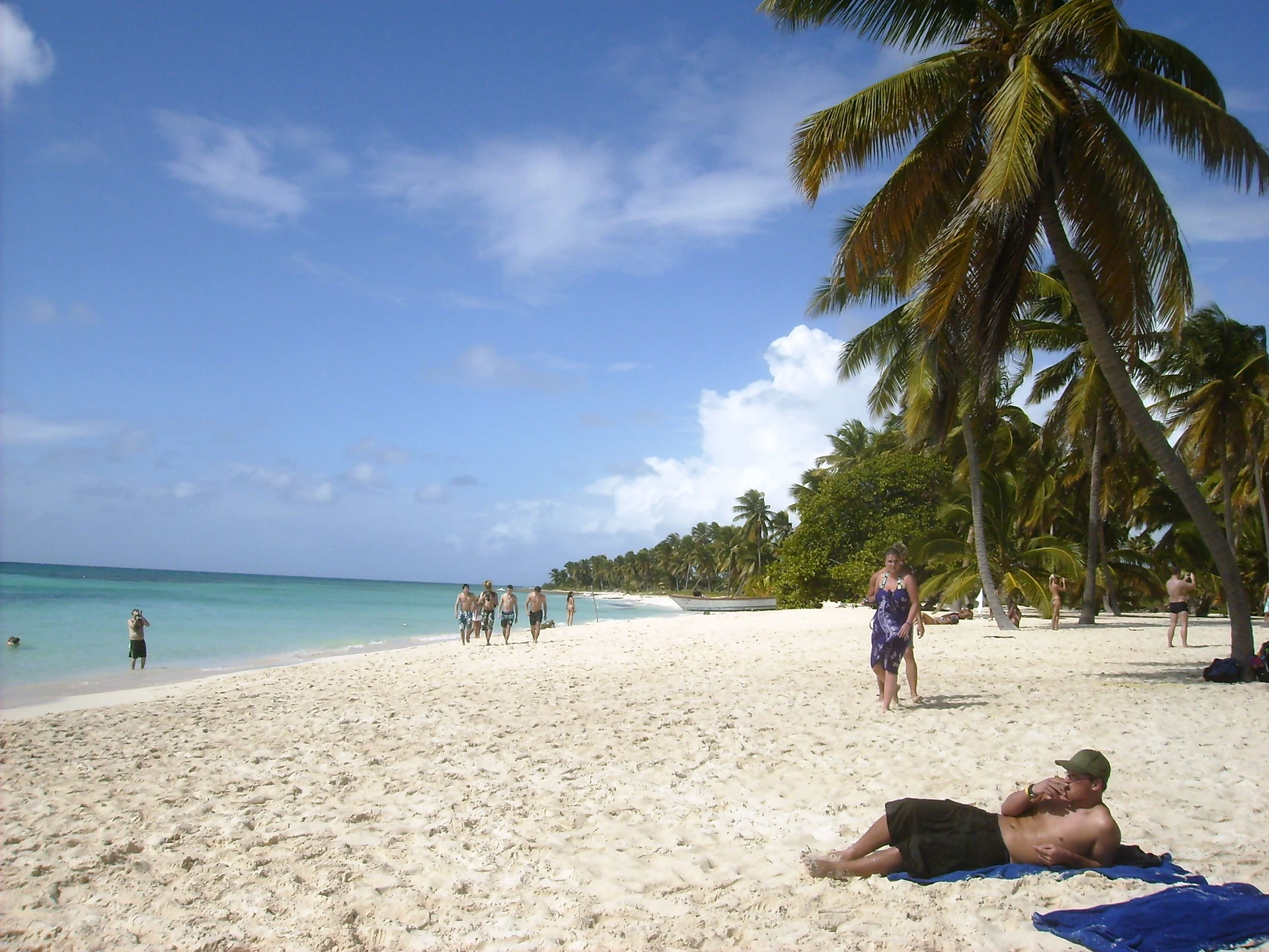 Visitantes caminham por uma ampla praia branca de Saona ladeada de coqueiros, junto à água turquesa calma das Caraíbas.