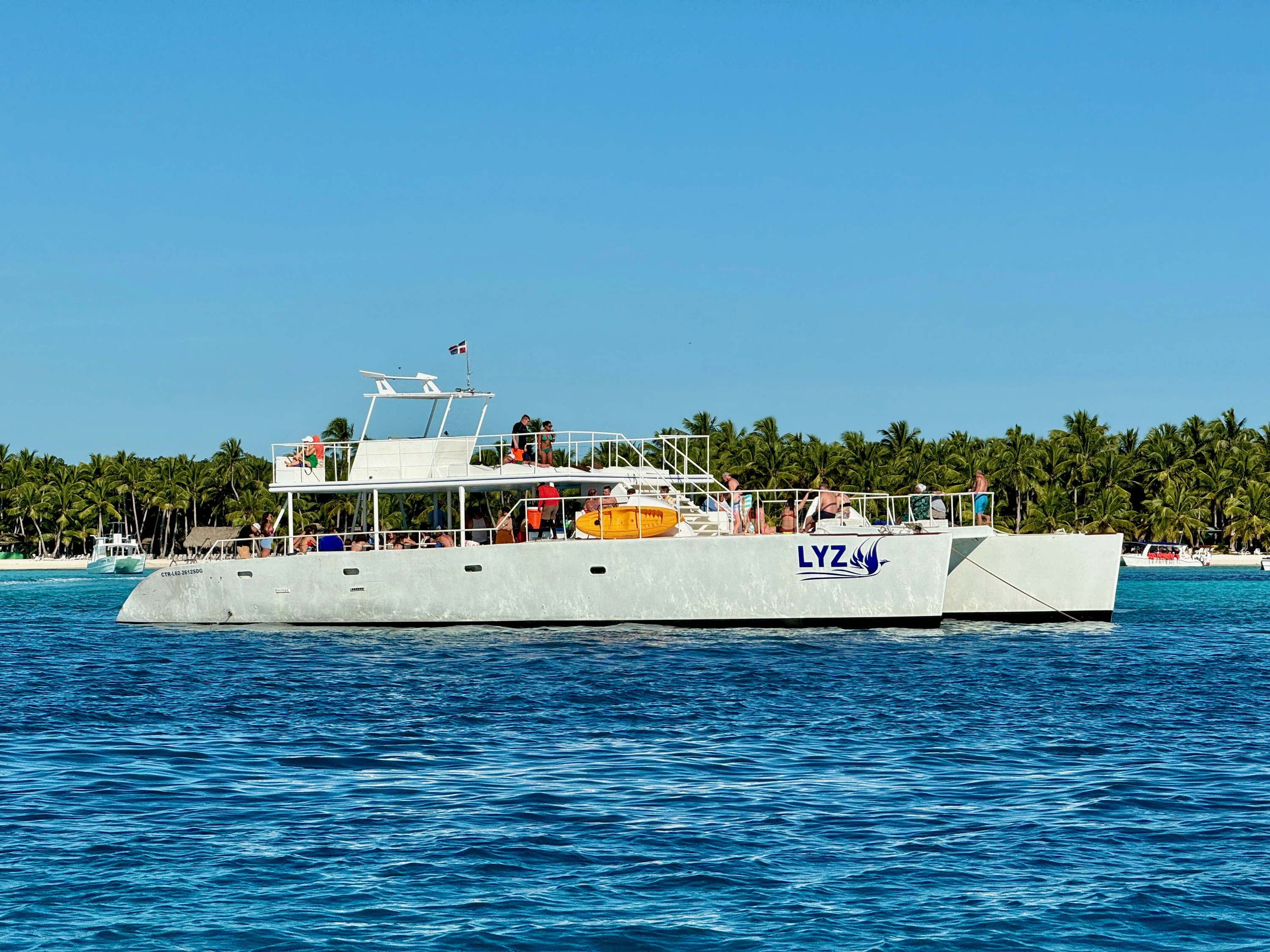 Un grande catamarano bianco da escursione galleggia in acque blu al largo della spiaggia di Saona bordata di palme.
