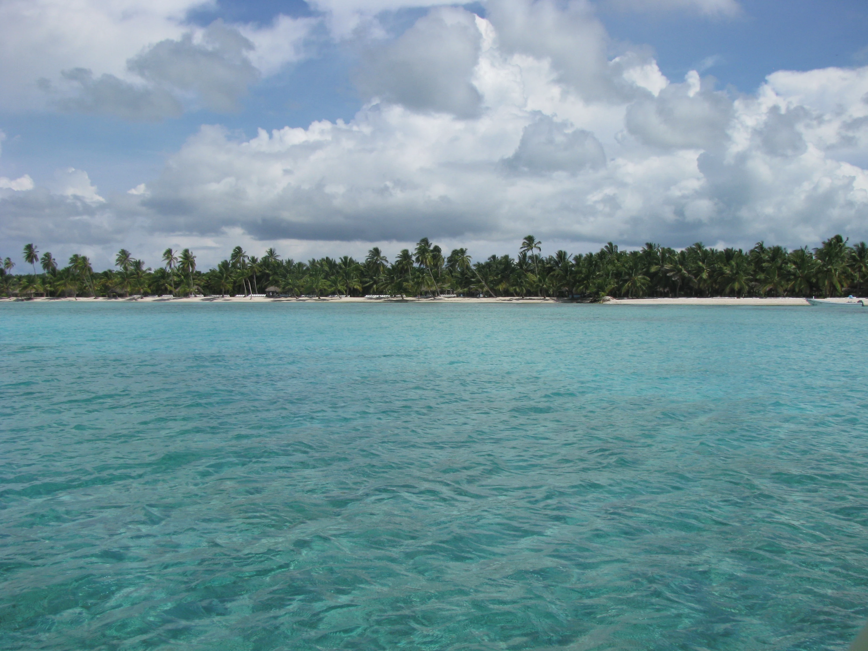 Acque turchesi si estendono verso la costa di sabbia bianca e le palme di Saona sotto grandi nuvole.