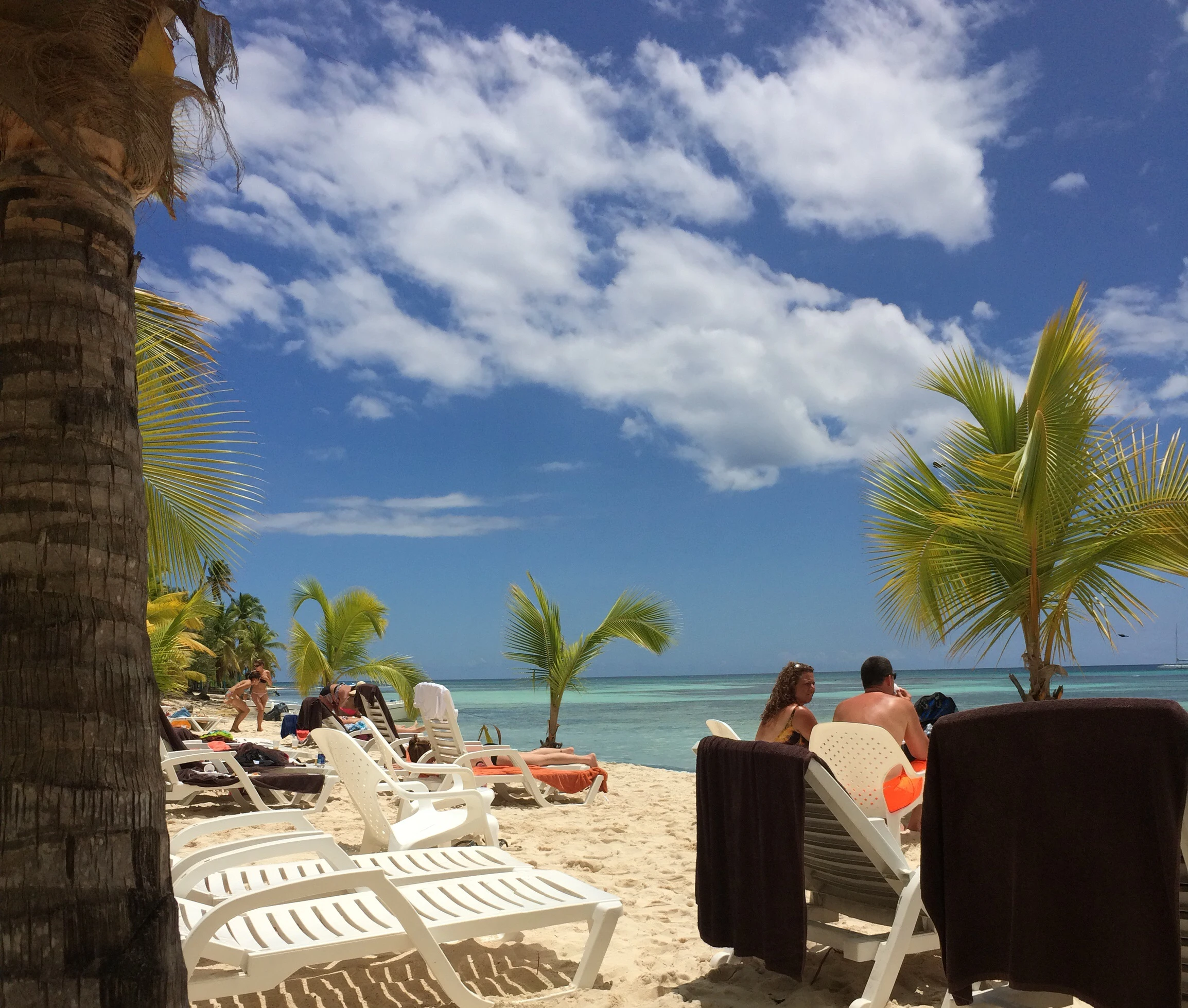 Besucher entspannen auf Stühlen im Palmenschatten neben einem weißen Saona-Strand und klarem blaugrünem Wasser.
