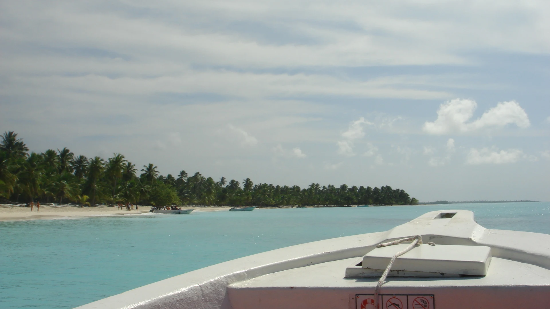 La prua di una barca punta verso la sabbia chiara e la costa di Saona bordata di palme, oltre acque turchesi.