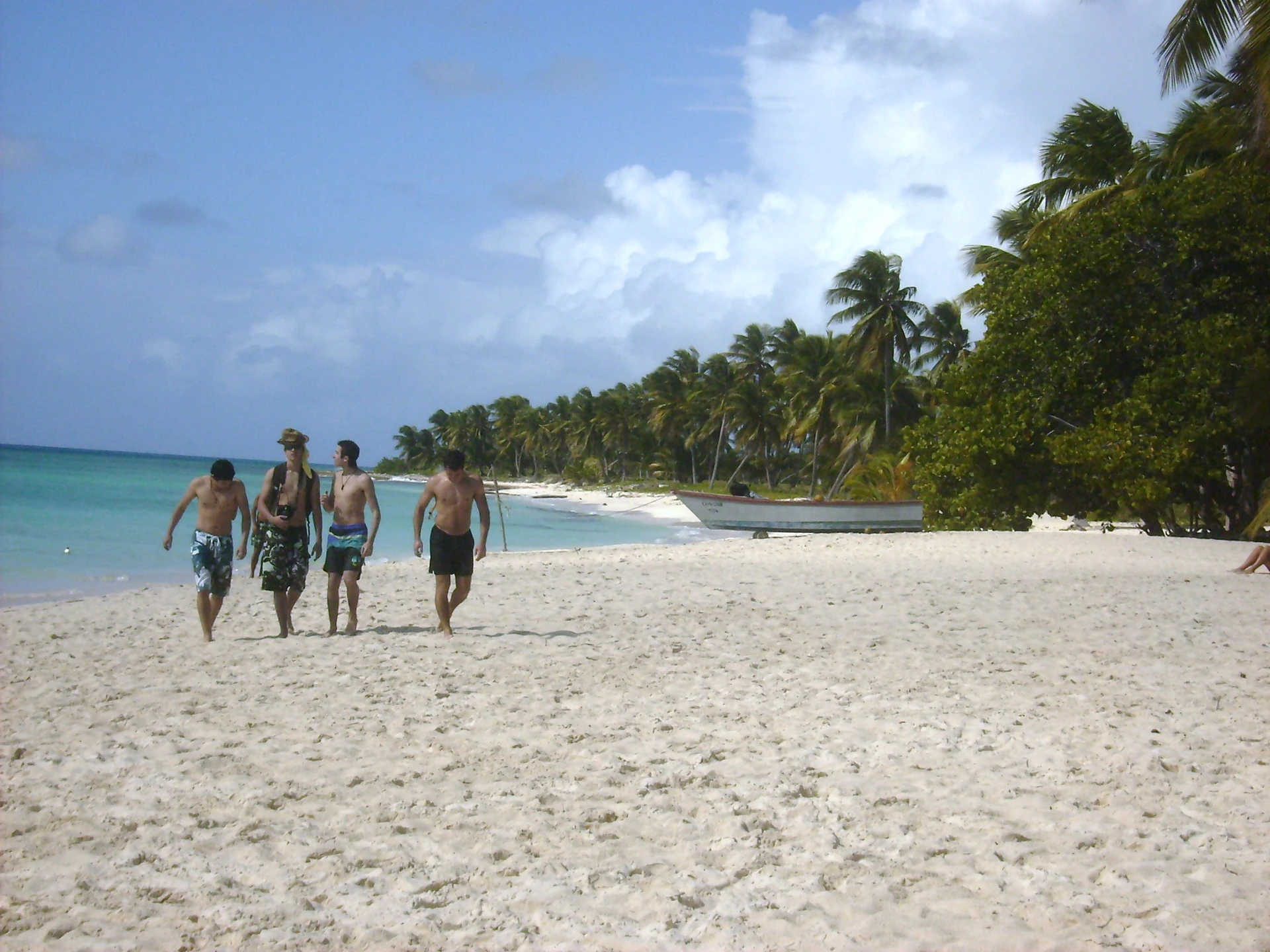 I visitatori camminano lungo una spiaggia bianca di Saona, accanto ad acqua turchese, palme fitte e una piccola barca in legno.
